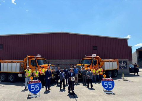 Photo of Illinois Governor Pritzker at a news conference in front of a small crowd, red building and two orange dump trucks